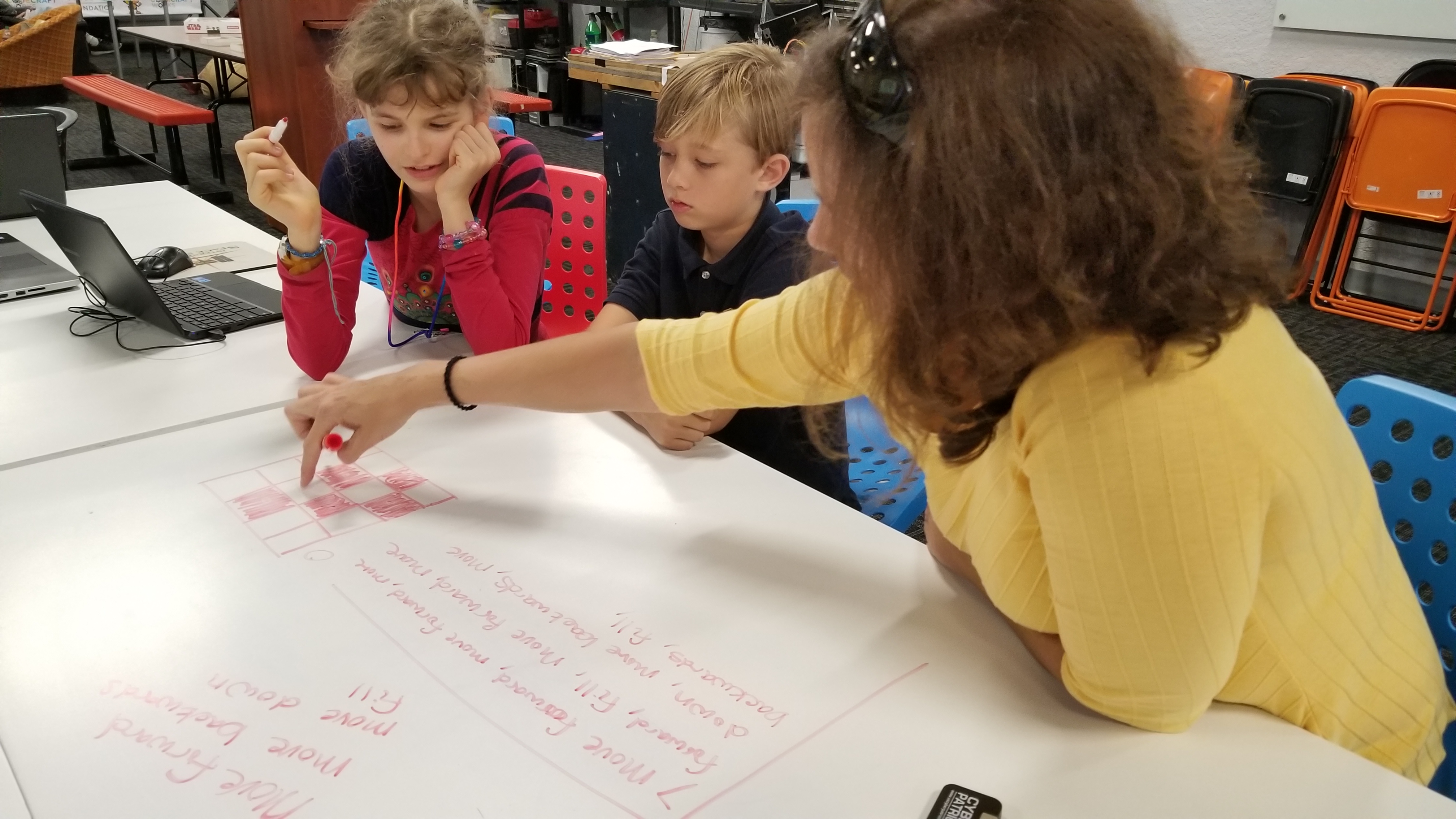 A woman in a yellow shirt points at a grid of colored squares drawn on a white table. A young girl in a red and black shirt and a boy in a black polo look on.