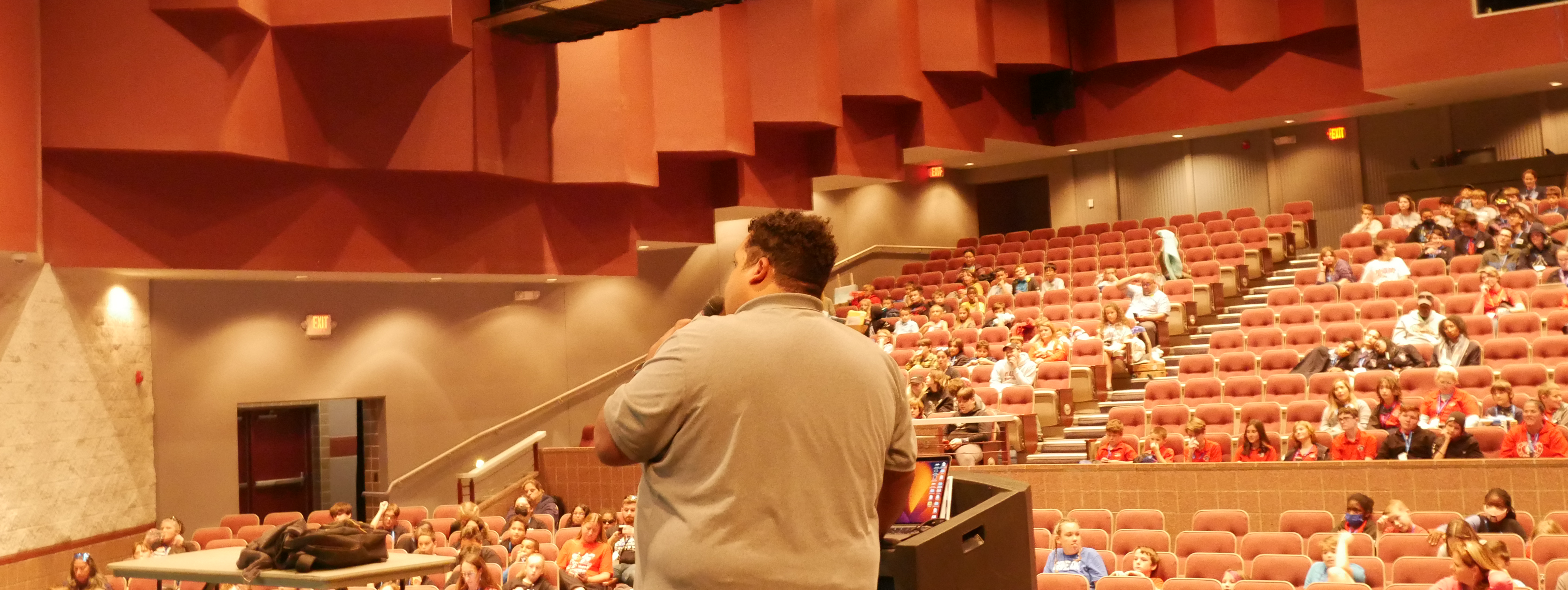 A photo of the back of a man speaking into a microphone, standing at a podium, and facing people seated in an auditorium.