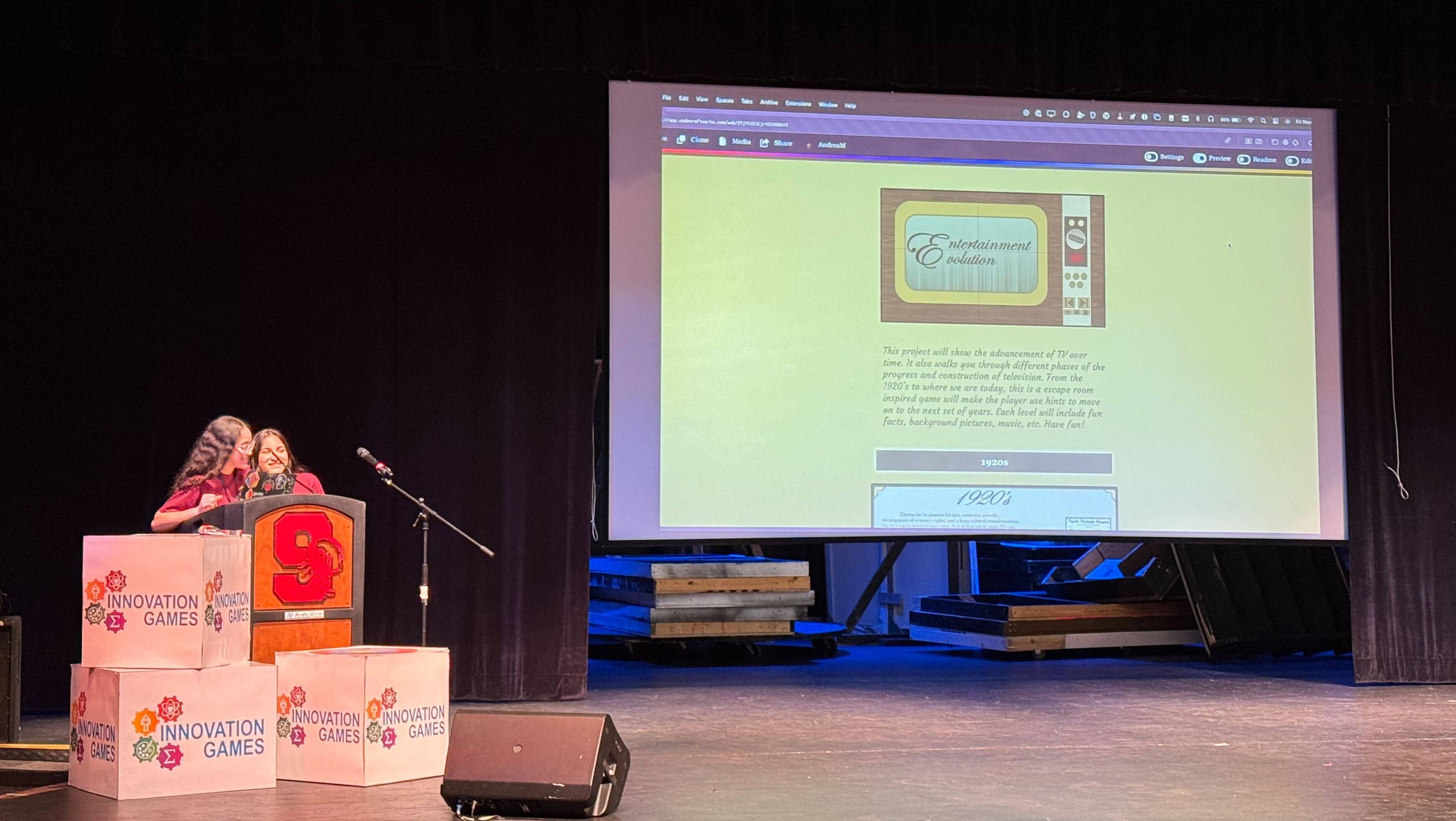 A photo of two girls at a podium on the left side of a stage, with a website shown on a projector screen above the center of the stage.