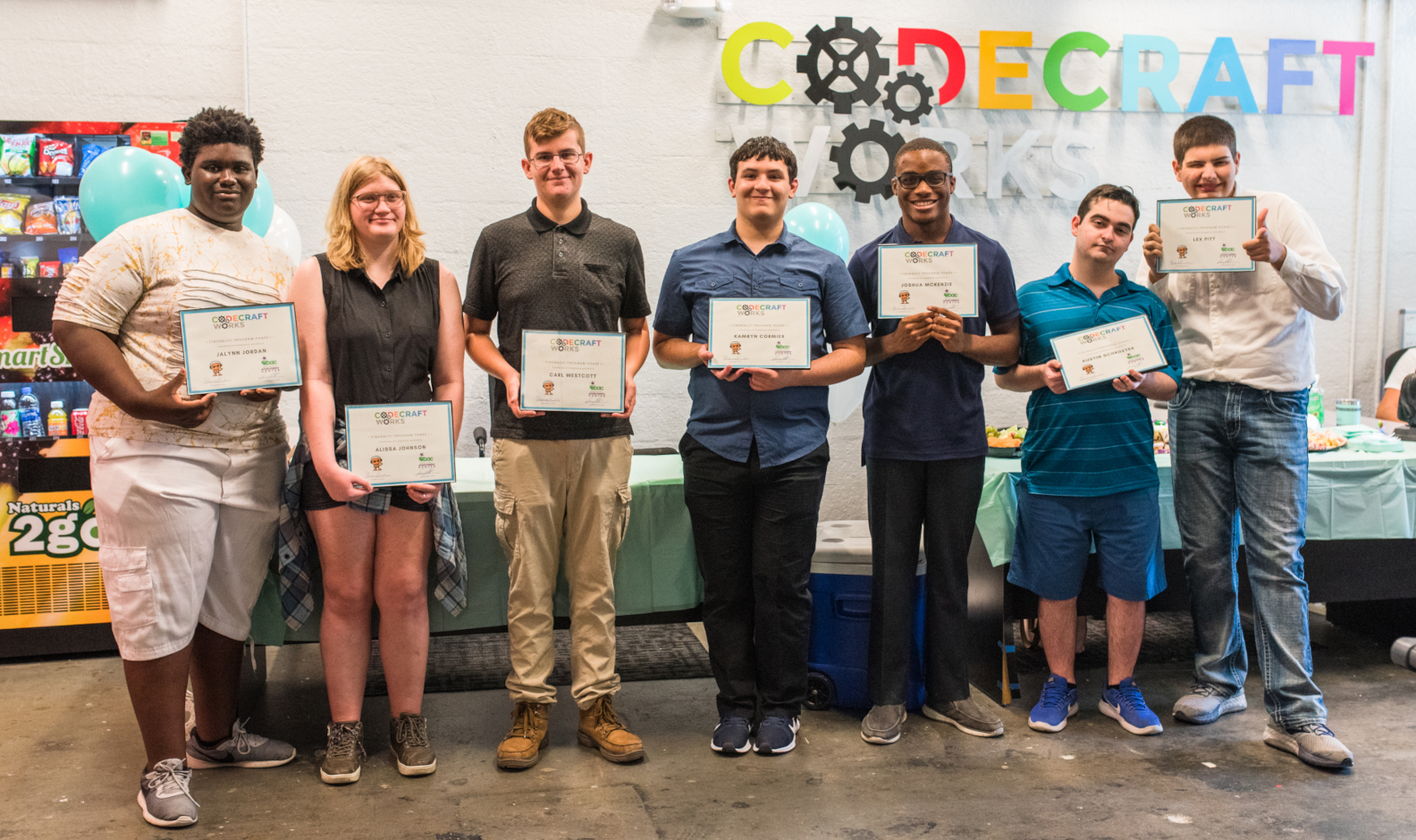 A photo of seven smiling students holding diplomas in a room with a large sign that reads 'Codecraft Works' on the wall behind them.