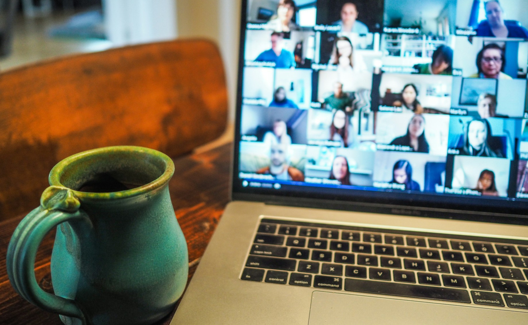 A photo of a laptop and a mug resting on a wooden table. The laptop screen displays a video conference with tiles of different peoples' video feeds taking up the whole screen.